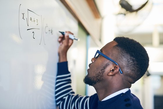 An African American man in glasses writing a mathmatical formula on a white board.