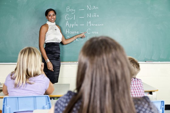 A teacher in front of a chalkboard with Spanish-English translations and students listening to her.